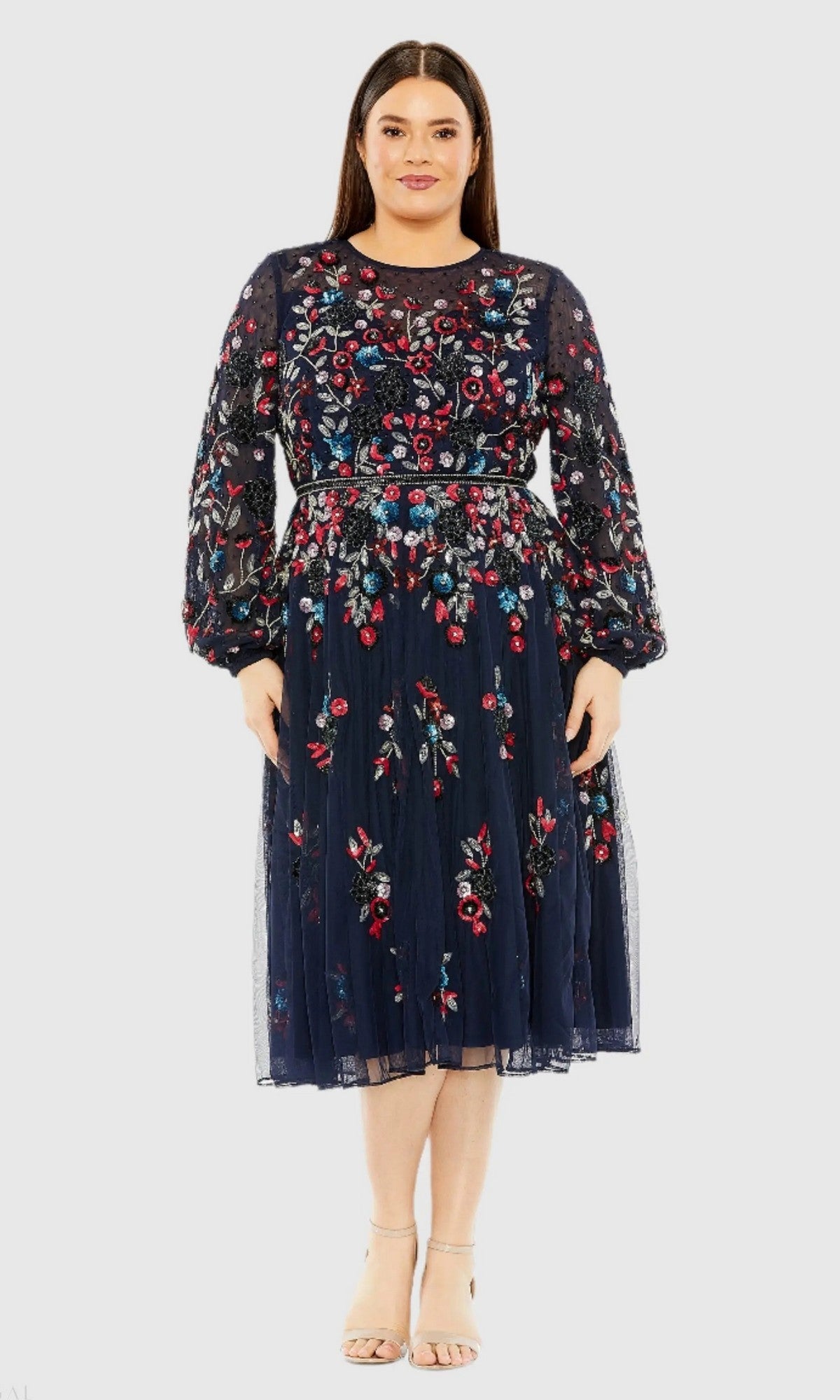 A woman stands facing forward, wearing the Mac Duggal 94050 Plus-Size Cocktail Dress with Sleeves in Midnight—a knee-length dress adorned with colorful floral beadwork—paired with beige heels.